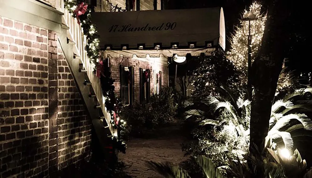 The image shows a warmly lit brick-clad entryway to a building named 17 Hundred 90 adorned with festive decorations and flanked by green plants creating a charming and inviting nighttime scene