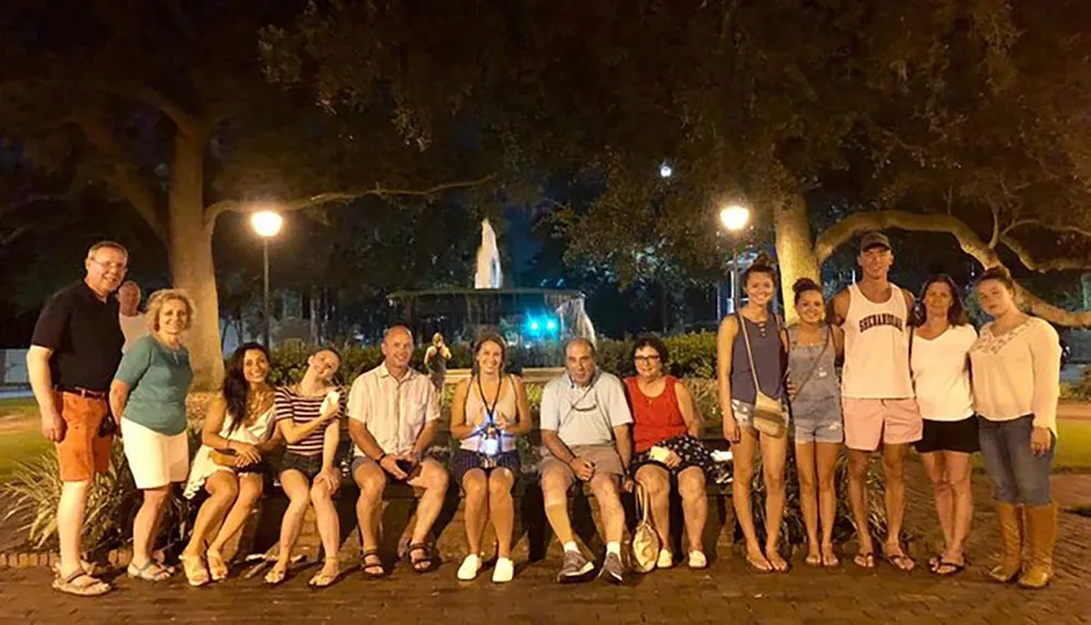 A group of smiling people is posing for a photo at night in an outdoor setting with trees and a fountain illuminated in the background