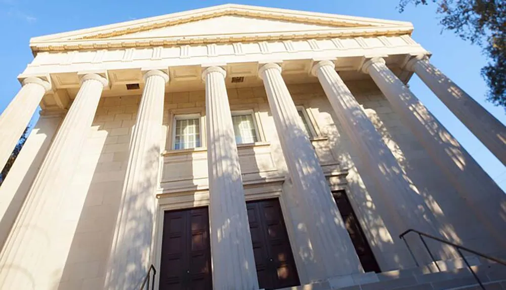 The image shows the facade of a neoclassical building characterized by its large columns and grand entrance with steps leading up to a set of tall wooden doors