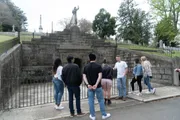 A group of people are standing near a fenced-off statue in what appears to be a cemetery.
