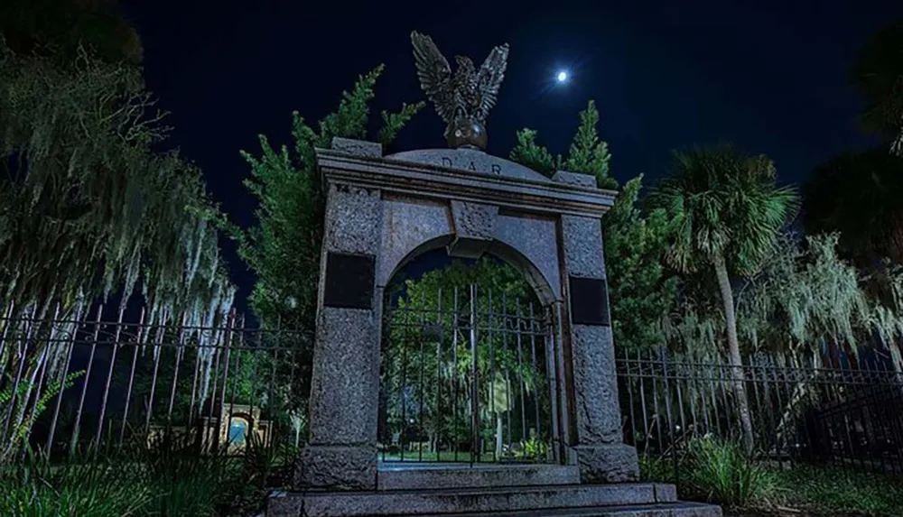 The image depicts a moonlit night scene with a grand ornate metal gate topped by an eagle statue surrounded by an atmosphere of gothic intrigue created by overhanging Spanish moss and silhouetted trees