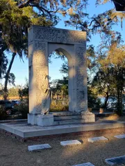 The image displays an ornate stone monument featuring a central archway with intricate carvings and a draped statue within, surrounded by several headstones, set against a backdrop of Spanish moss-draped trees and a clear sky.