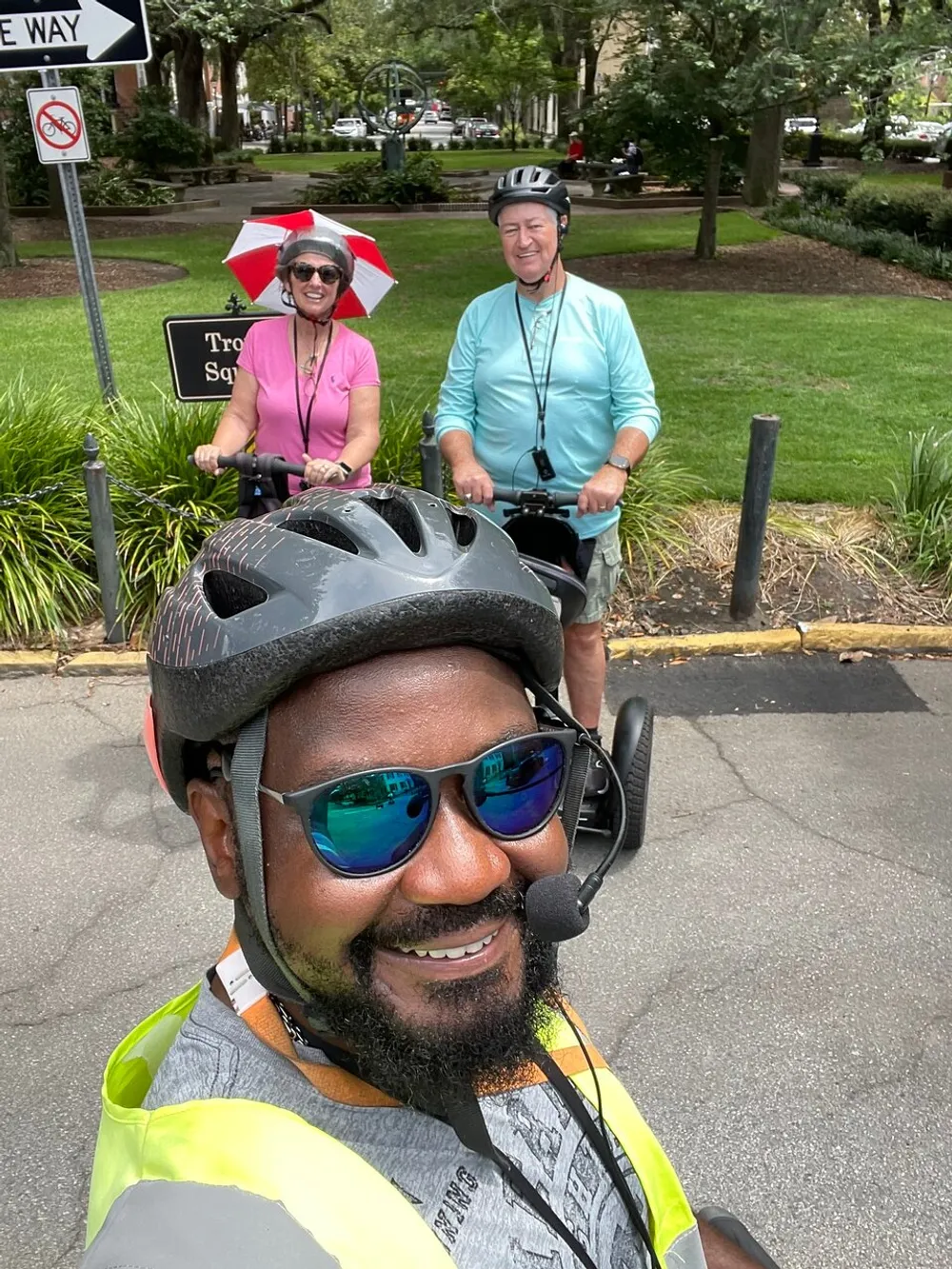 Three people are smiling for a selfie during a bicycle ride with one person wearing a helmet with a camera and the other two in the background one of whom is using a hat with an umbrella attachment