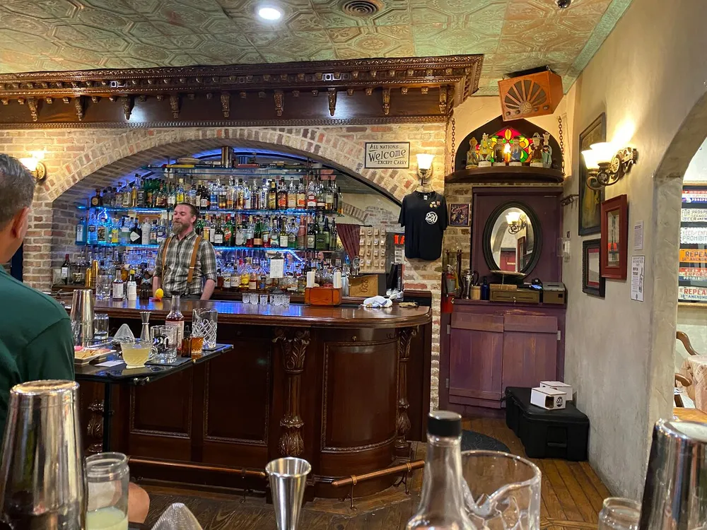 The image shows the interior of a cozy well-decorated bar with a bartender standing behind the counter surrounded by an assortment of bottles and bar paraphernalia