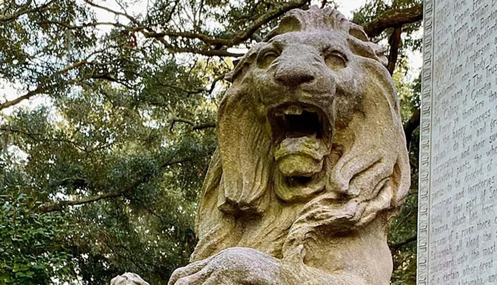 The image shows a stone sculpture of a roaring lion next to an engraved monument or tablet set against a backdrop of green foliage