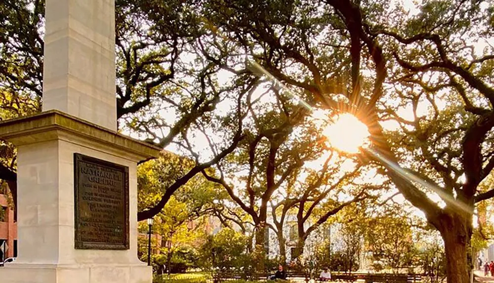 The image captures a serene park scene with the sun beaming through the branches of grand oak trees highlighting a monument with a commemorative plaque and people enjoying the outdoors in the background