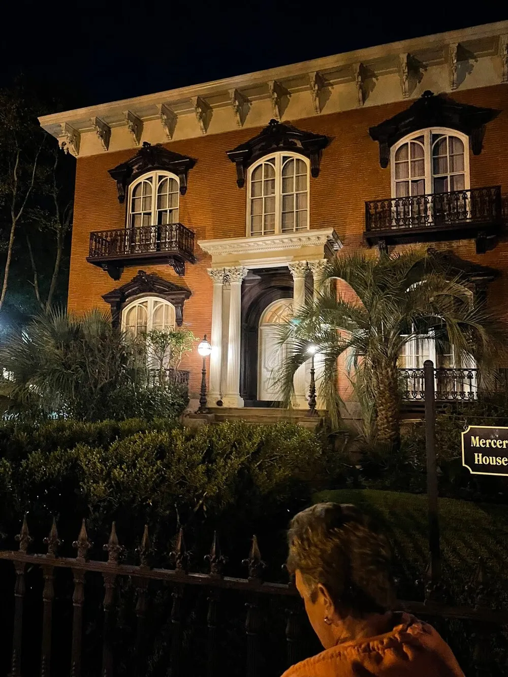 The image shows an illuminated historic brick building at night viewed by a person standing in the foreground with ornate balconies and a sign indicating it is the Mercer House