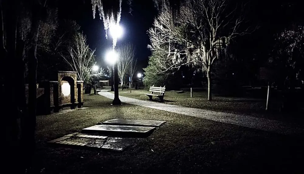 A dimly lit empty park at nighttime with benches and a pathway illuminated by street lamps