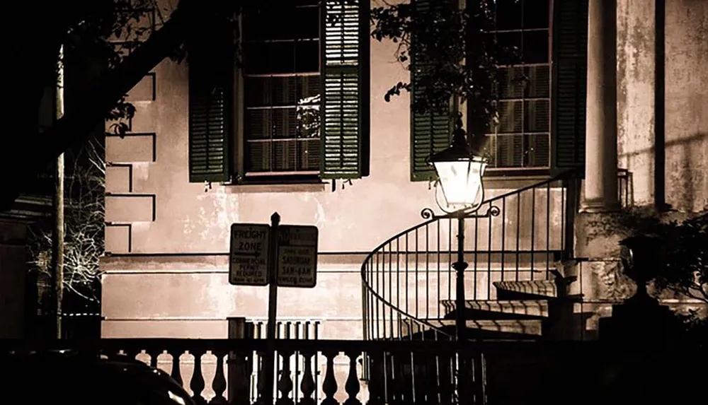 A sepia-toned image captures a tranquil night scene featuring street signs a lit lantern and the exterior of a building with shutters and a staircase railing