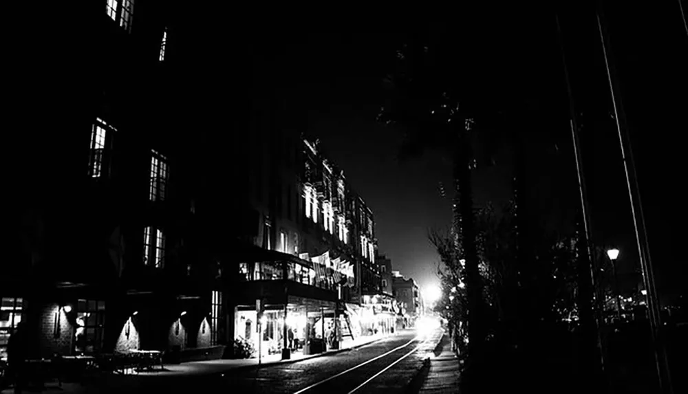 The image shows a nighttime street scene captured in black and white with illuminated windows and streetlights contrasting against the dark silhouettes of buildings and trees