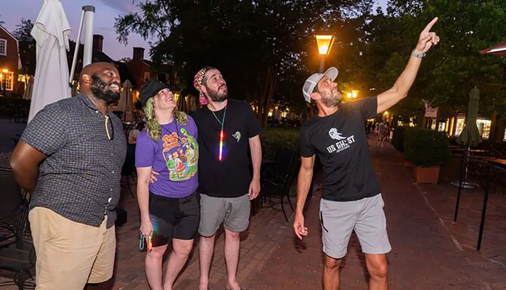 Four people are looking up with one person pointing towards the sky seemingly at something interesting or surprising in a public outdoor setting during the evening