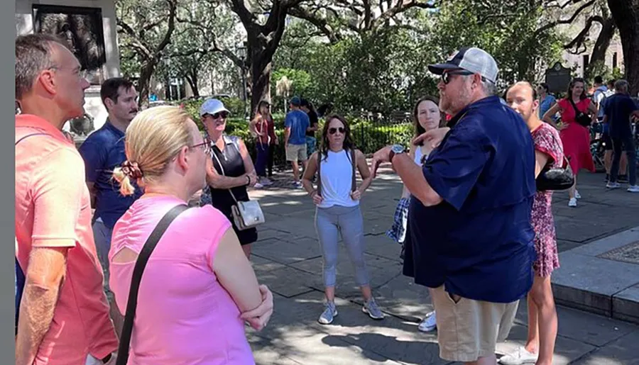 A group of people is attentively listening to a tour guide in an outdoor setting with trees in the background.
