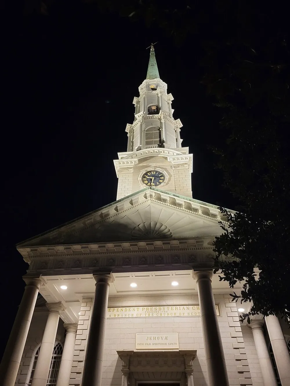 The image shows the illuminated facade of a church at night with a prominent steeple and classical architecture including columns and a pediment with inscriptions