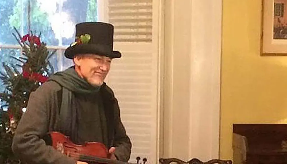 A person wearing a top hat adorned with festive ornaments is smiling and holding a ukulele in a room decorated for Christmas