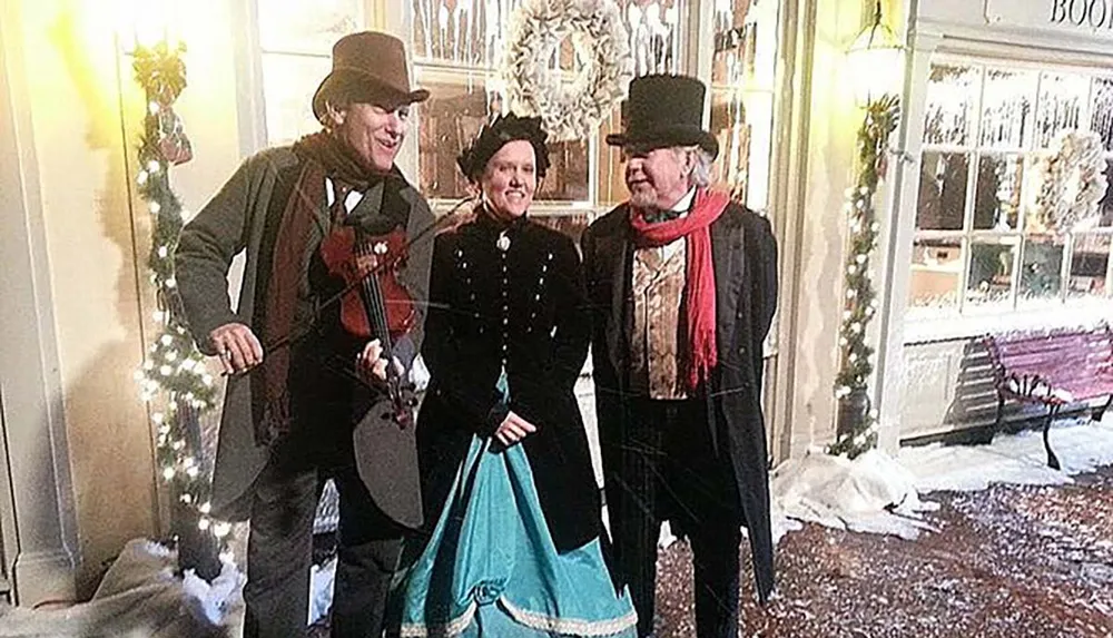Three individuals are dressed in historical costumes exuding a festive spirit with one playing a violin amidst a decorated snowy setting reminiscent of a Charles Dickens novel
