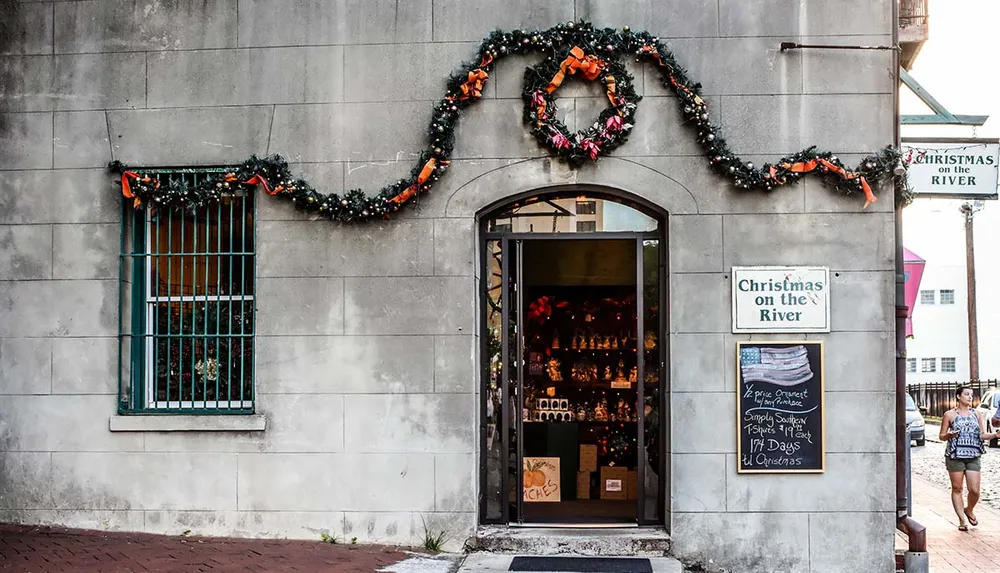 A festive storefront is adorned with holiday decorations including garland and a large wreath with signs indicating it is Christmas on the River and featuring holiday-themed promotions