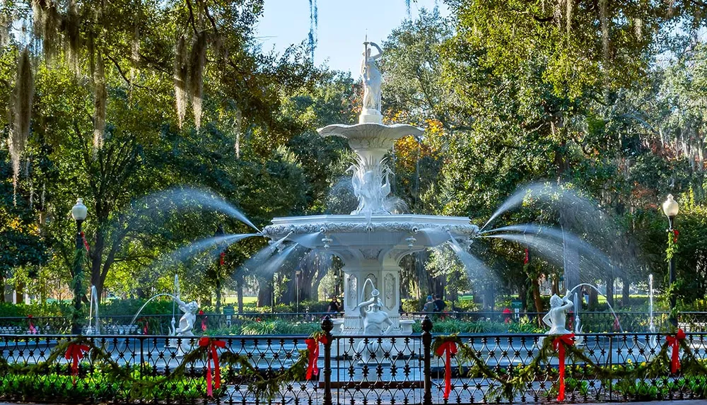 This is an ornate white fountain with water jets surrounded by a fence with red bows set against a backdrop of lush green trees