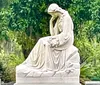 The image shows a marble statue of a contemplative figure seated with one hand to the face set against a backdrop of lush greenery