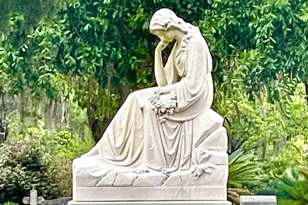 The image shows a marble statue of a contemplative figure seated with one hand to the face set against a backdrop of lush greenery