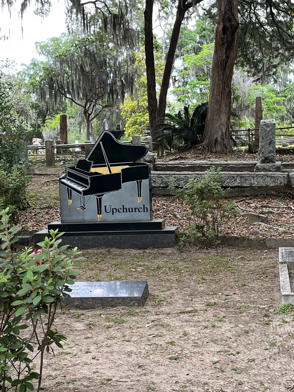 The image features a unique piano-shaped headstone with the name Upchurch in a peaceful tree-laden cemetery