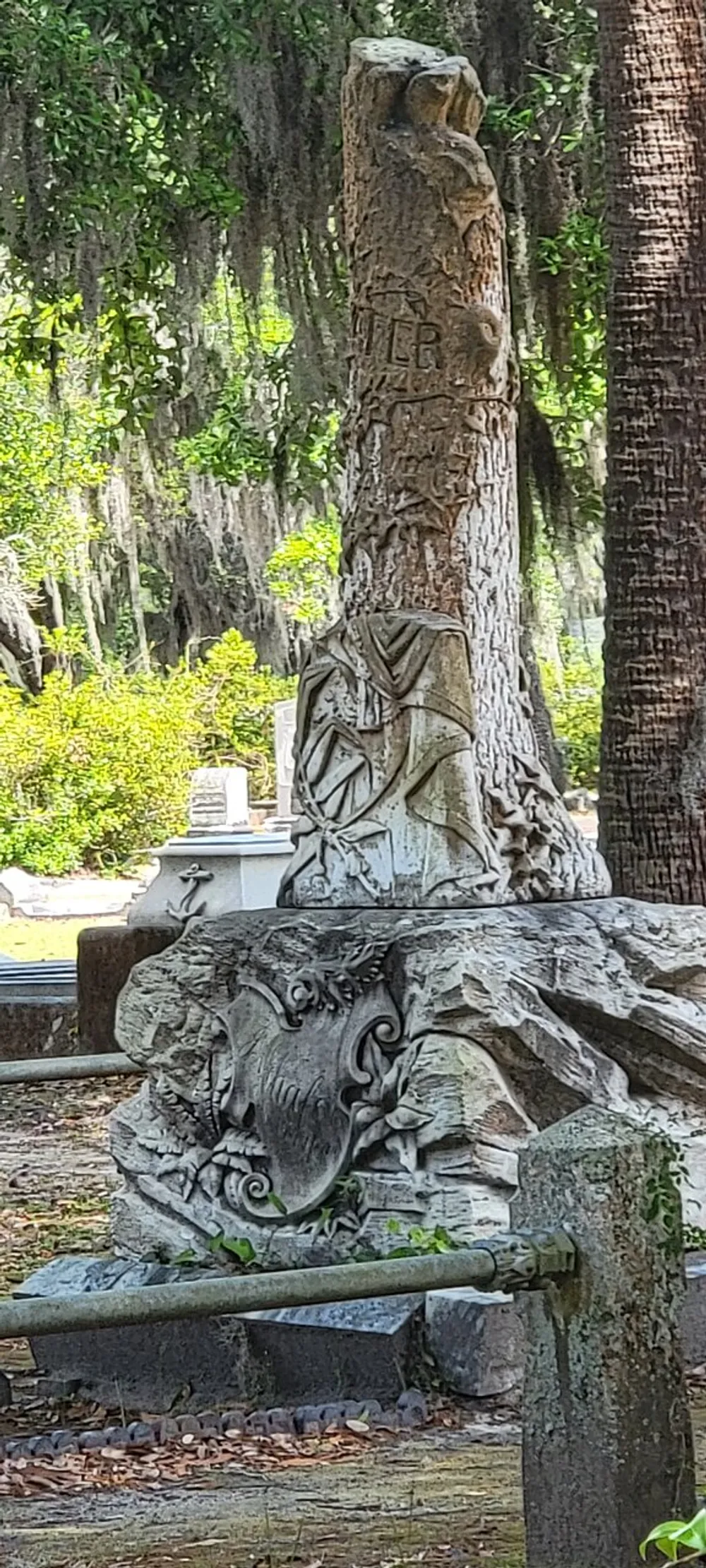 The image shows a weathered stone sculpture possibly part of a grave or monument featuring a draped figure and decorative elements set against a backdrop of trees draped with Spanish moss