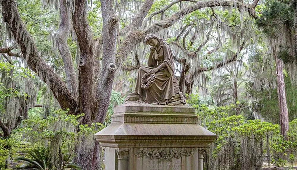 A solemn statue of a seated figure with a wreath rests atop a pedestal surrounded by the ethereal draping of Spanish moss in a serene wooded area