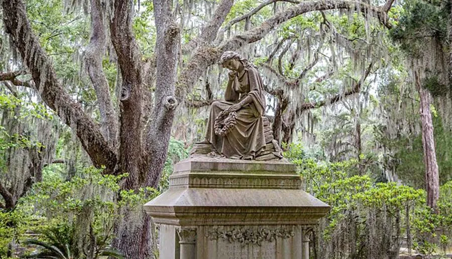 A solemn statue of a seated figure with a wreath rests atop a pedestal surrounded by the ethereal draping of Spanish moss in a serene, wooded area.