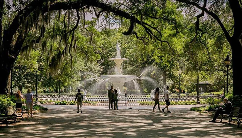 The image features a serene park setting where people are enjoying a sunny day around a grand fountain shaded by large trees with hanging moss
