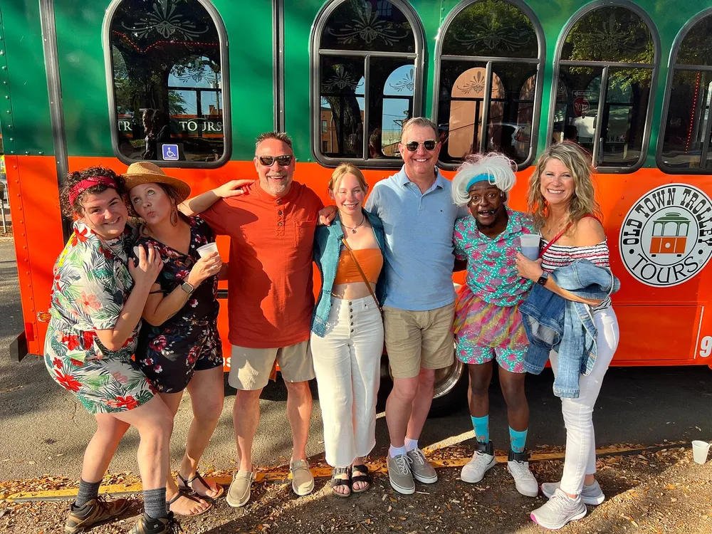 A cheerful group of people poses for a photo in front of an orange Old Town Trolley Tours trolley bus with some participants playfully interacting and smiling at the camera