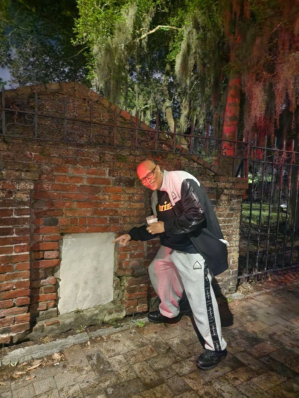A person wearing glasses casual clothes and a cap is pointing at a bricked-up section in a wall while holding a beverage in the other hand against a backdrop of Spanish moss-draped trees and iron fence in what looks like an evening setting