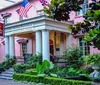 The image shows a charming and historic pink building with pillars and a sign indicating The Olde Pink House Restaurant  Tavern surrounded by green foliage and flying American and Georgian flags