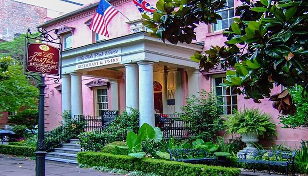 The image shows a charming and historic pink building with pillars and a sign indicating The Olde Pink House Restaurant  Tavern surrounded by green foliage and flying American and Georgian flags