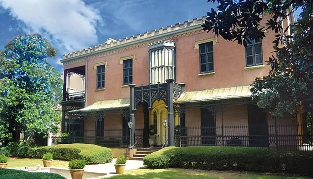 The image shows an elegant two-story pink stucco building with black ironwork detailing on the balconies and porch set against a backdrop of greenery and a partly cloudy blue sky
