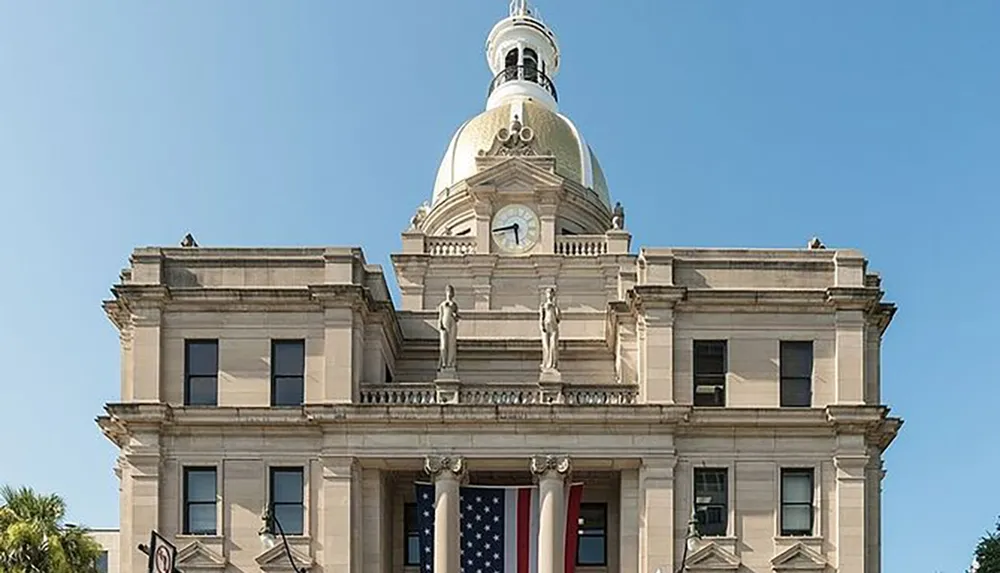 This image shows a grand historic building with a large American flag hanging from its facade characterized by classical architecture elements such as statues columns and a dome with a clock