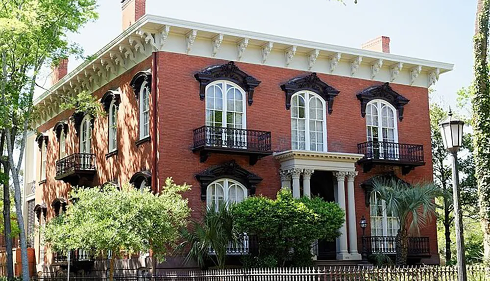 The image shows a two-story red brick house with ornate black balconies large arched windows a columned entrance and surrounded by green foliage