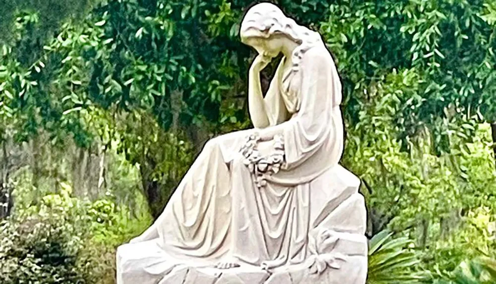 The image shows a white statue of a seated pensive female figure against a backdrop of lush green vegetation