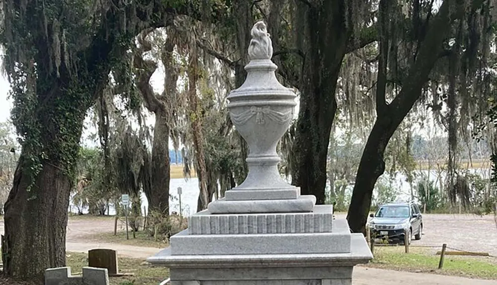 The image shows a large ornate urn-shaped stone monument in a cemetery with Spanish moss-covered trees and a vehicle in the background
