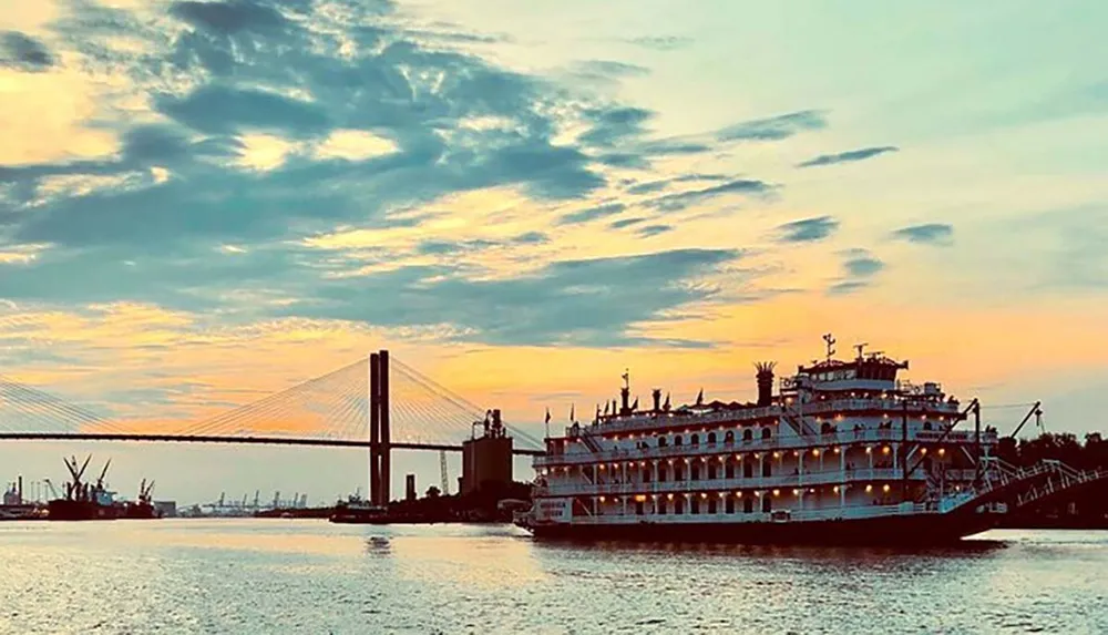 A paddle steamer is docked near a bridge against the backdrop of a colorful sunset sky