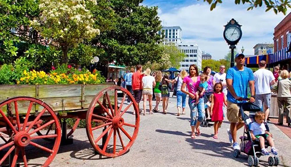 This image shows people strolling through a vibrant urban park area featuring a flower-filled wagon and a classic street clock suggesting a leisurely family-friendly atmosphere