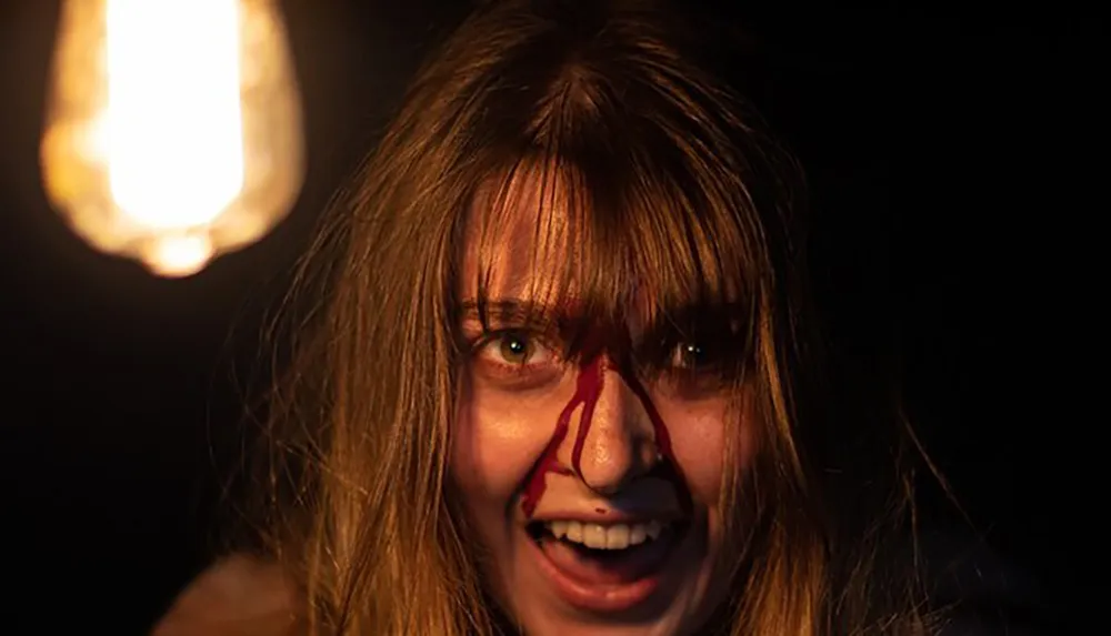 A person is depicted with a frenzied expression and a stream of blood running down their face set against a dark background with a blurred light source