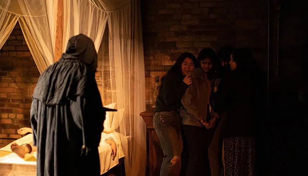 A figure in a dark cloak stands ominously near a bed with body parts as a group of apprehensive visitors watches in a dimly lit eerie setup suggesting a horror-themed attraction or theatrical performance