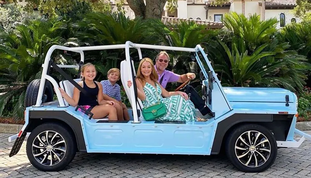 A family of four is smiling while posing in a light blue open-air vehicle with a tropical backdrop