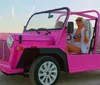 A woman in a white bikini is smiling while sitting in a pink open-top vehicle on a beach at sunset