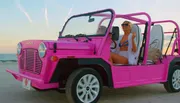 A woman in a white bikini is smiling while sitting in a pink open-top vehicle on a beach at sunset.
