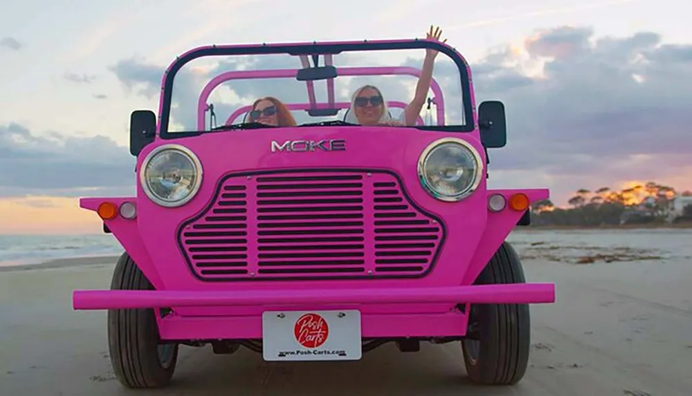 Two people are waving from a bright pink beach vehicle with a sunset in the background