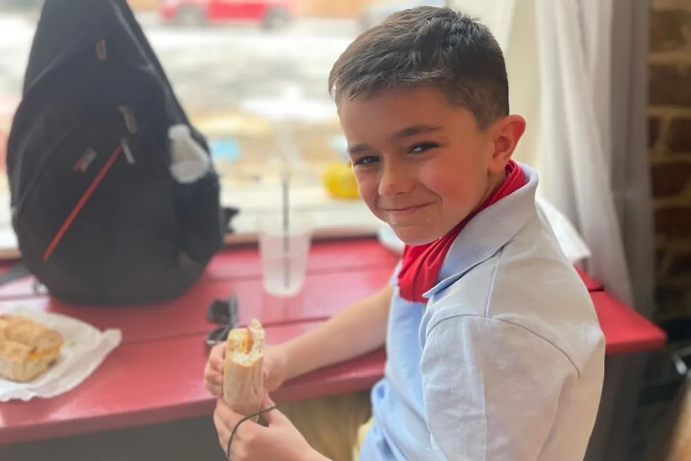 A smiling boy is sitting at a red table holding a half-eaten sandwich with a backpack behind him and a glass of water on the table in a room with a sunny window