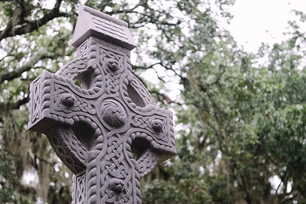 The image displays an intricately carved stone Celtic cross set against a blurred background of leafy trees