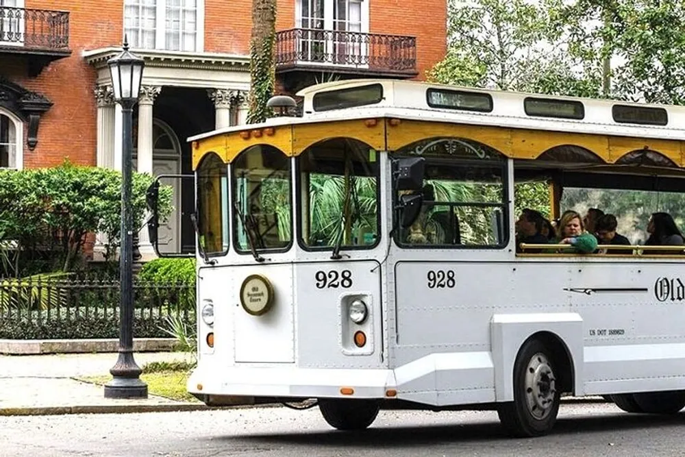 An old-fashioned trolley bus filled with passengers is driving by a leafy street with elegant buildings in the background