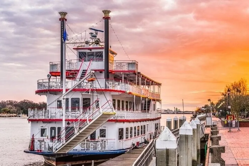 A traditional paddlewheel riverboat is docked along a riverwalk under a sunset sky