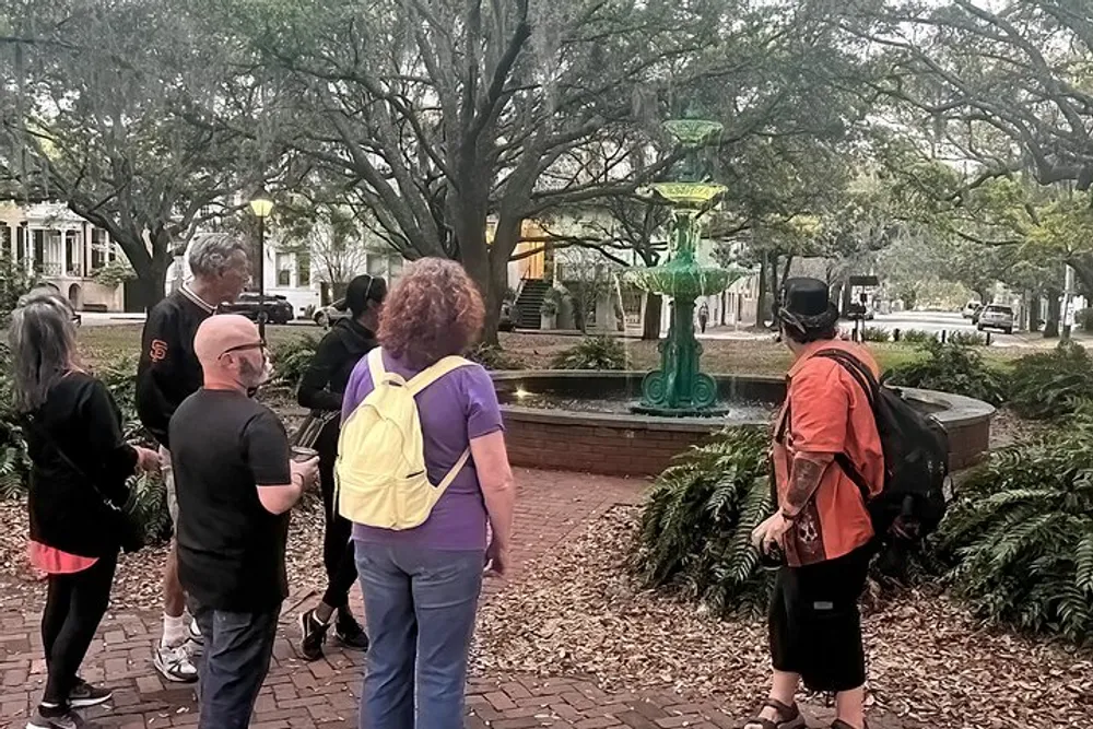 A group of people are gathered around a green fountain in a park with large trees and a residential street in the background